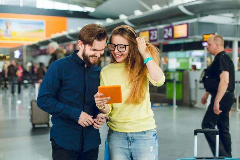 portrait-couple-standing-airport-she-has-long-hair-glasses-sweater-jeans-he-has-beard-shirt-pants-they-are-looking-tablet