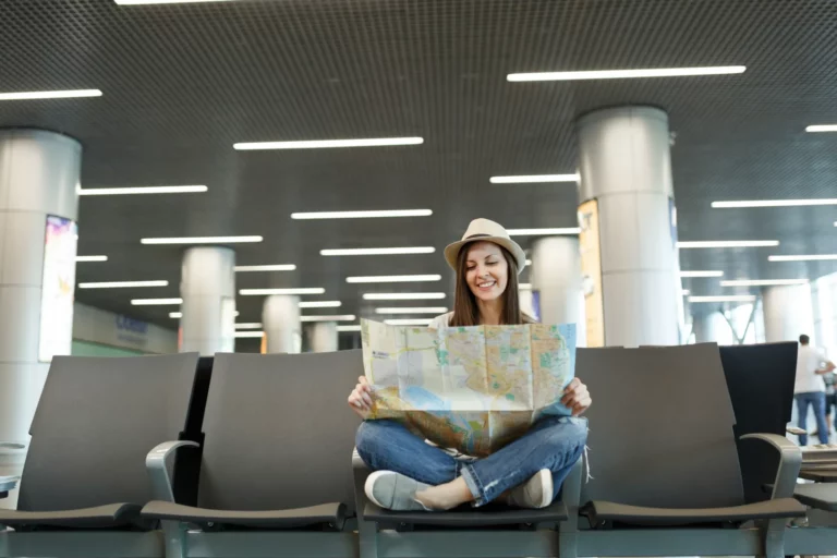 young-smiling-traveler-tourist-woman-with-crossed-legs-holding-paper-map-searching-route-waiting-lobby-hall-international-airport